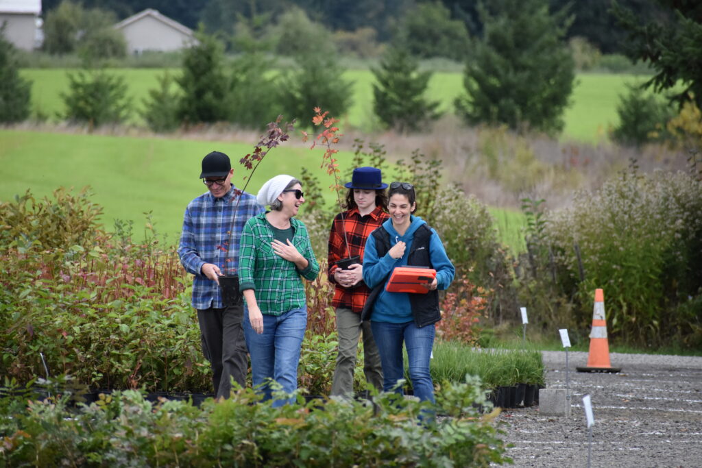 A Clean Water Services employee holds a clipboard while speaking to community members at a native plant giveaway located at the Tualatin River Farm. The community members hold native plants as they walk through the selection of more native plants.