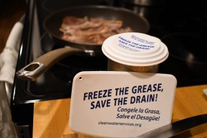 A pan of bacon sizzles in the background. The foreground shows a grease scraper leaning against a tin can with a grease lid from the 