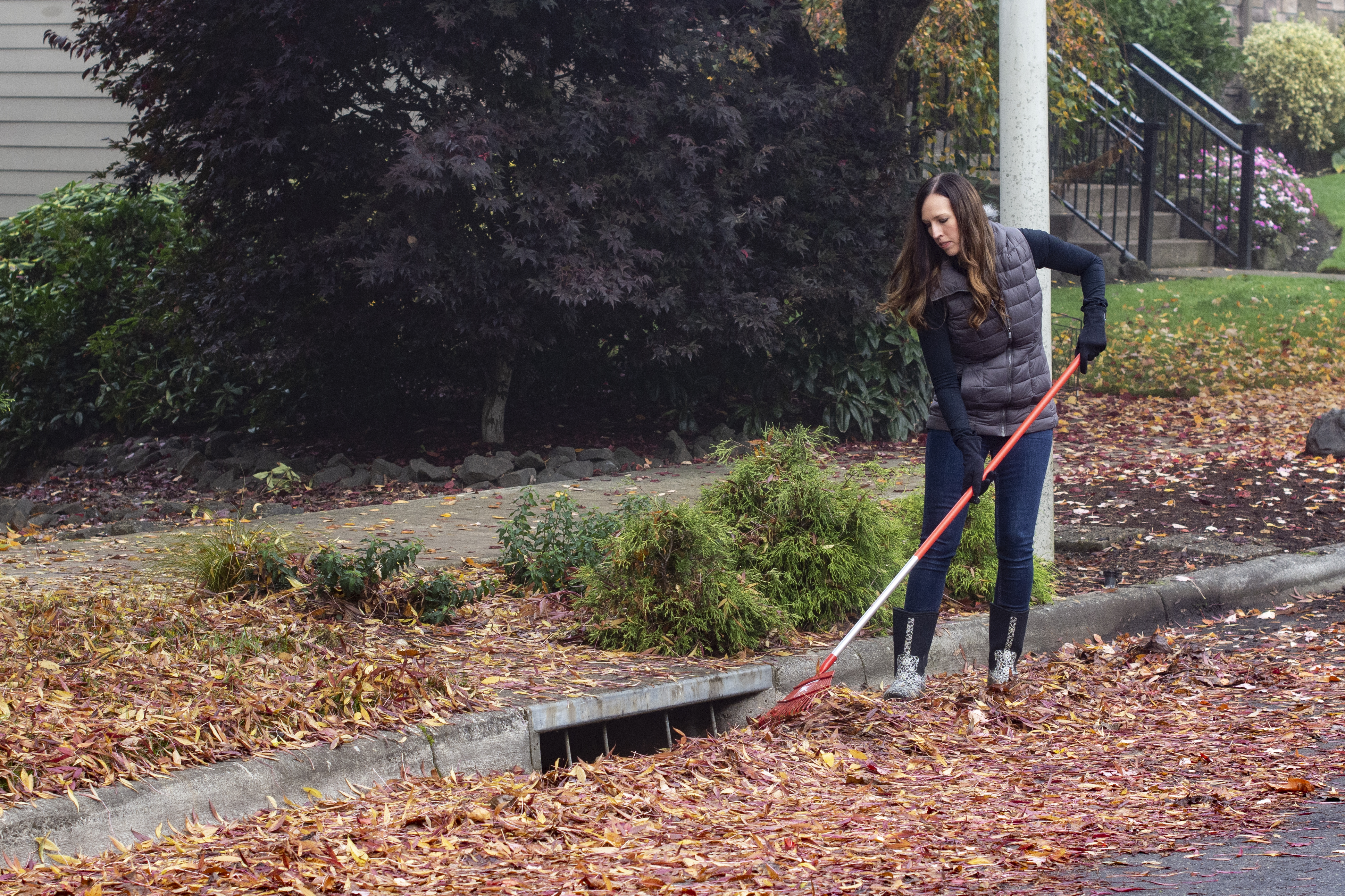 Leaves that have fallen off of a nearby tree cover a stormdrain. A community member works to rake the leaves away from the drain with a red rake while wearing a puff vest and rain boots.
