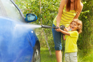 A Mother and Child wearing yellow shirts use a hose to wash off a blue car while parked on the grass.