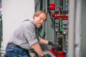 A Clean Water Services Employee works on an electric panel while in a uniform and smiling at the camera