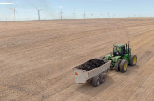 Green tractor pulling a large container of biosolids across farmland with wind turbines twirl in the background