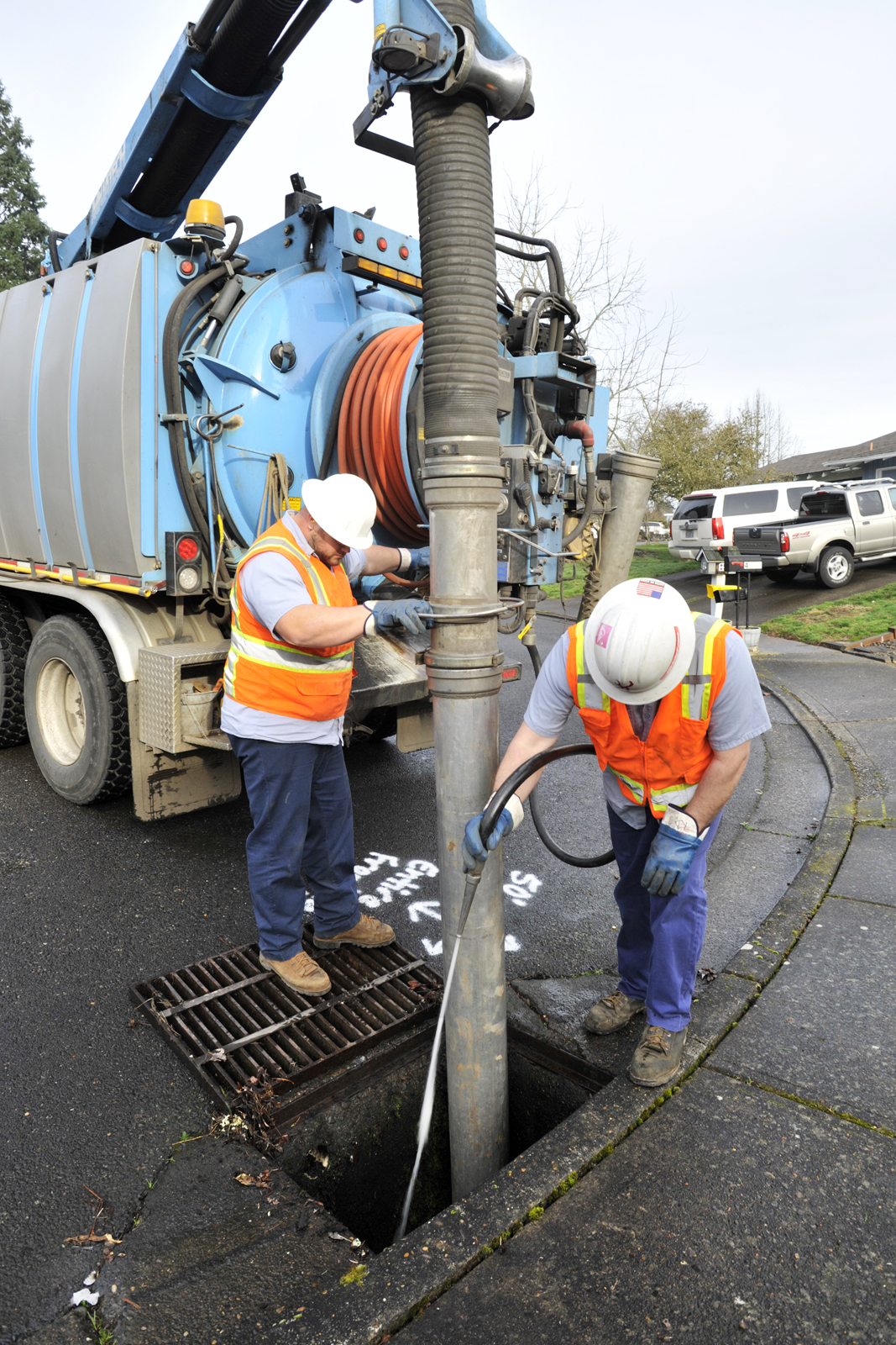 Two clean water services employees in hard hats and safety vests work with a vac truck to clean out a stormdrain.