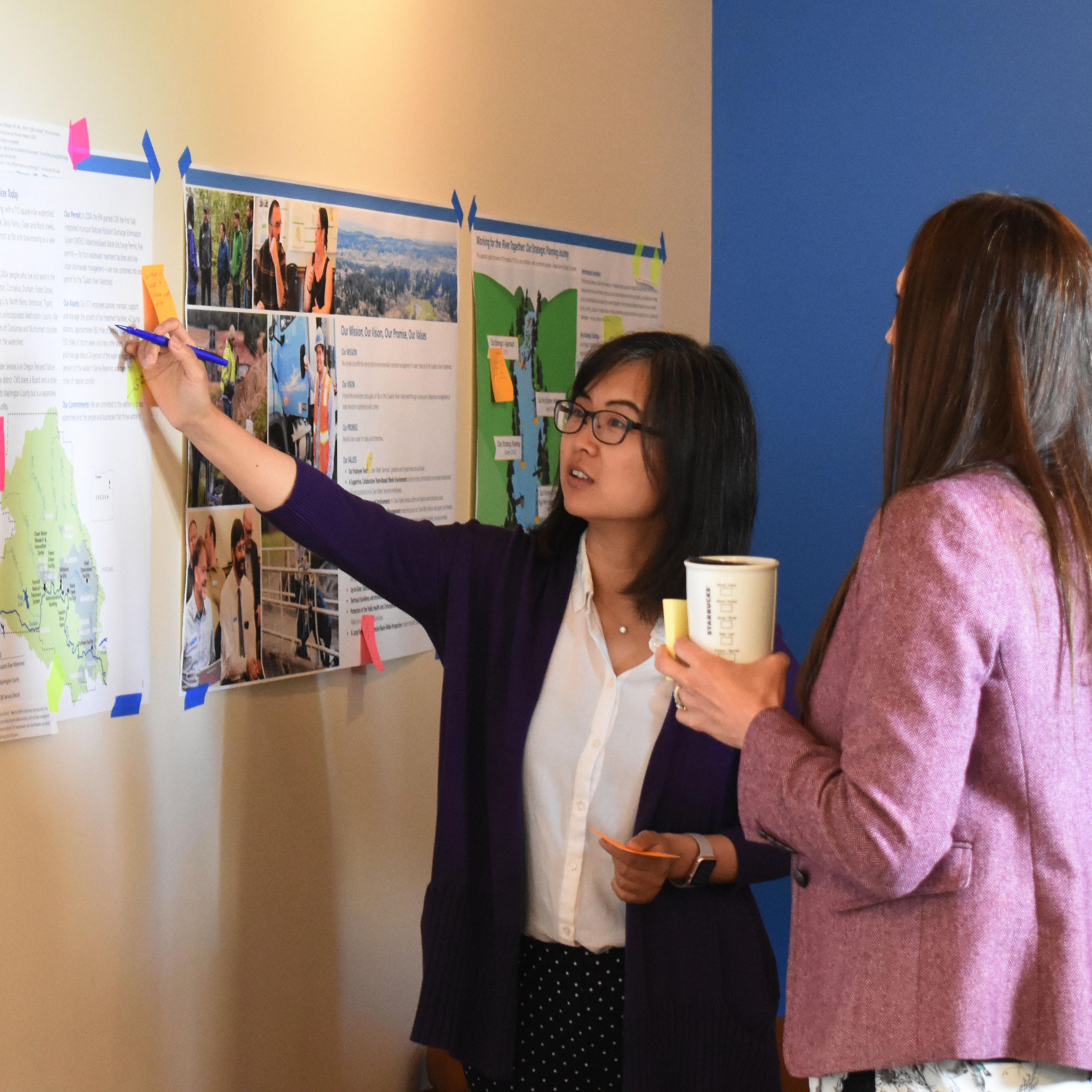Clean Water Services Employees talk and point to posters in a meeting room. Clean Water Services Employees talk and point to posters in a meeting room. ABC, Administrative Building Complex, Holly Dober, Jessica Owen, Strategic Approach, Ting Lu, meeting, Human, Person