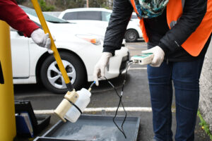 A Clean Water Services employee uses technology to test the water at an industry. the employee wears an orange vest and rubber gloves.