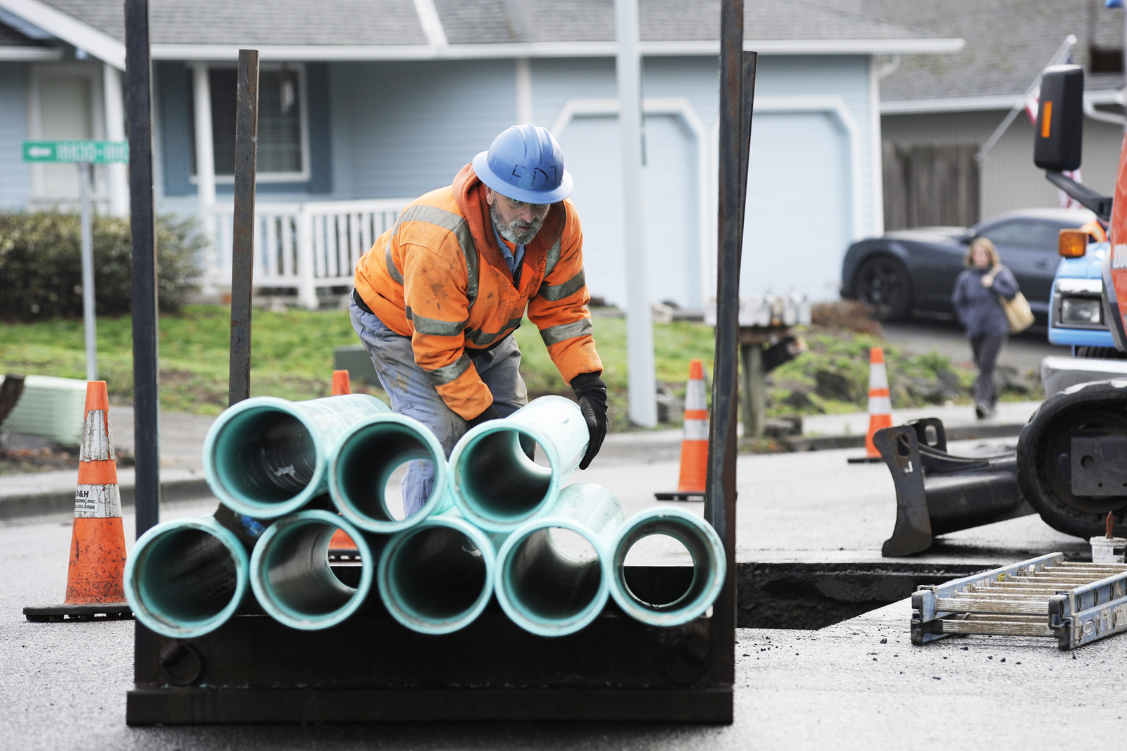 A Clean Water Services employee unloads pipes from a pile while wearing a hard hat and an orange safety jacket. He is in a Washington county neighborhood working on installation.
