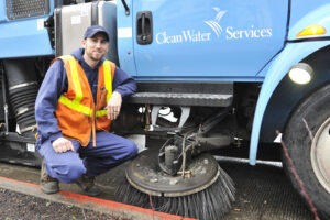 A Clean Water Services employee in an orange vest smiles as he kneels next to a Street Sweeper truck, showing off the brushes that clean the roads.