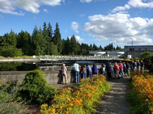 A large group of community members look into a clarifier at Clean Water Services' Rock Creek Water Resource Recovery Facility while on a Facility Tour. The blue sky and green trees contrast the orange flowers lining the walkway to the clarifier.