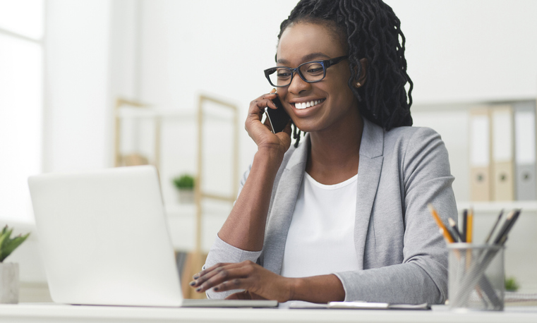 Woman working on a computer while on the phone. she smiles while wearing a gray blazer.