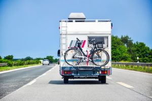 a camper RV drives down a road with a bicycle on the back on a sunny day.