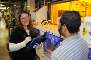 A Clean Water Services employee speaks with an industry worker while on an inspection. They stand inside the industry's building and the Inspector holds a clipboard while wearing PPE.