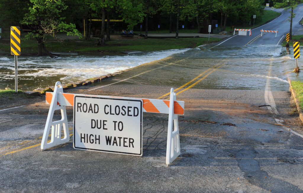 A barricade sign reading "Road Closed Due to High Water" sits on a road in front of a flooded area of road.