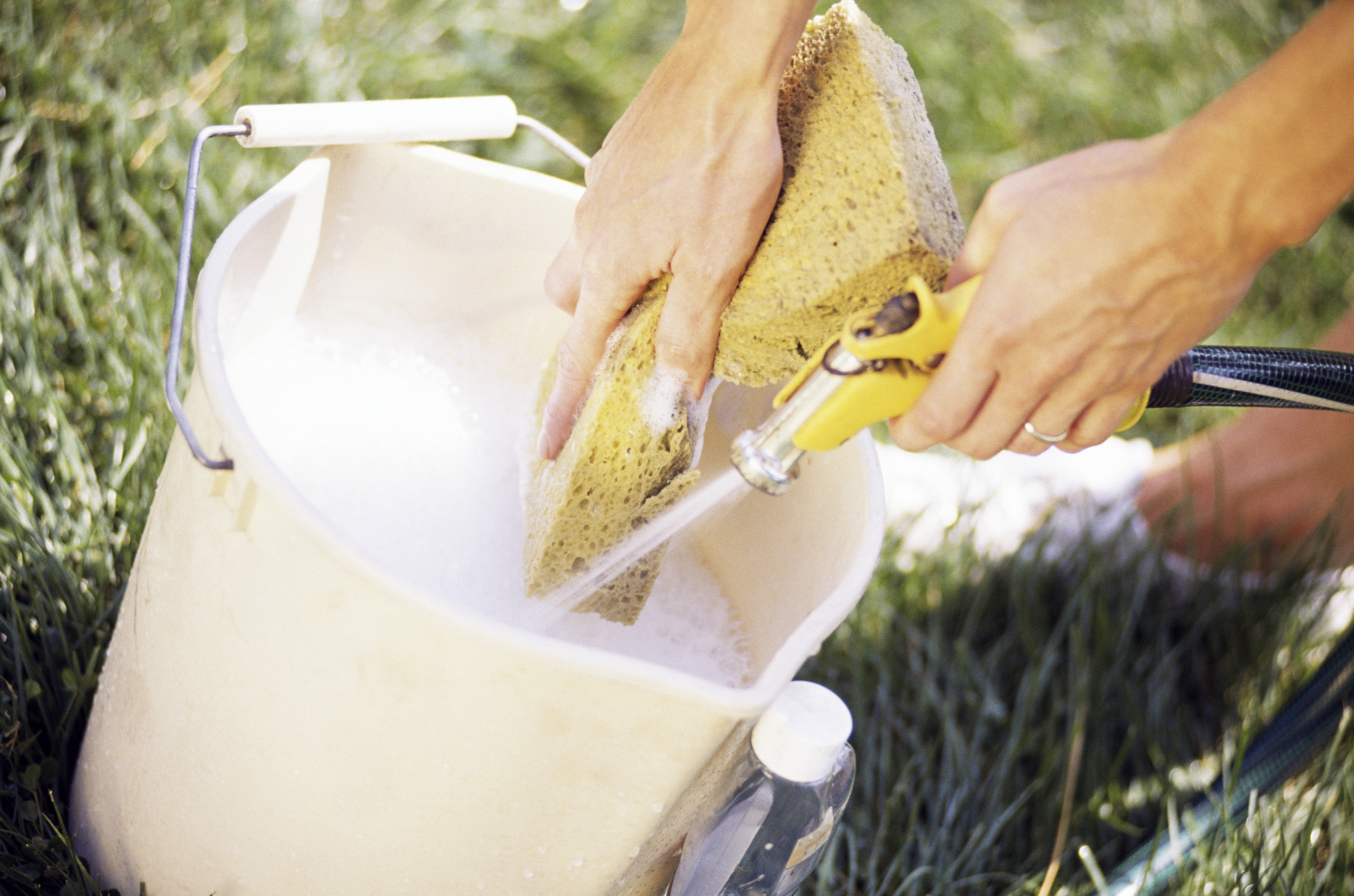 Soapy hands hold a sponge and a hose while filling up a bucket with soapy water in preparation to wash a car. The bucket sits on the grass with a small bottle of soap.
