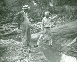 Old photo of two men with one standing across the Tualatin, one foot on each river bank.