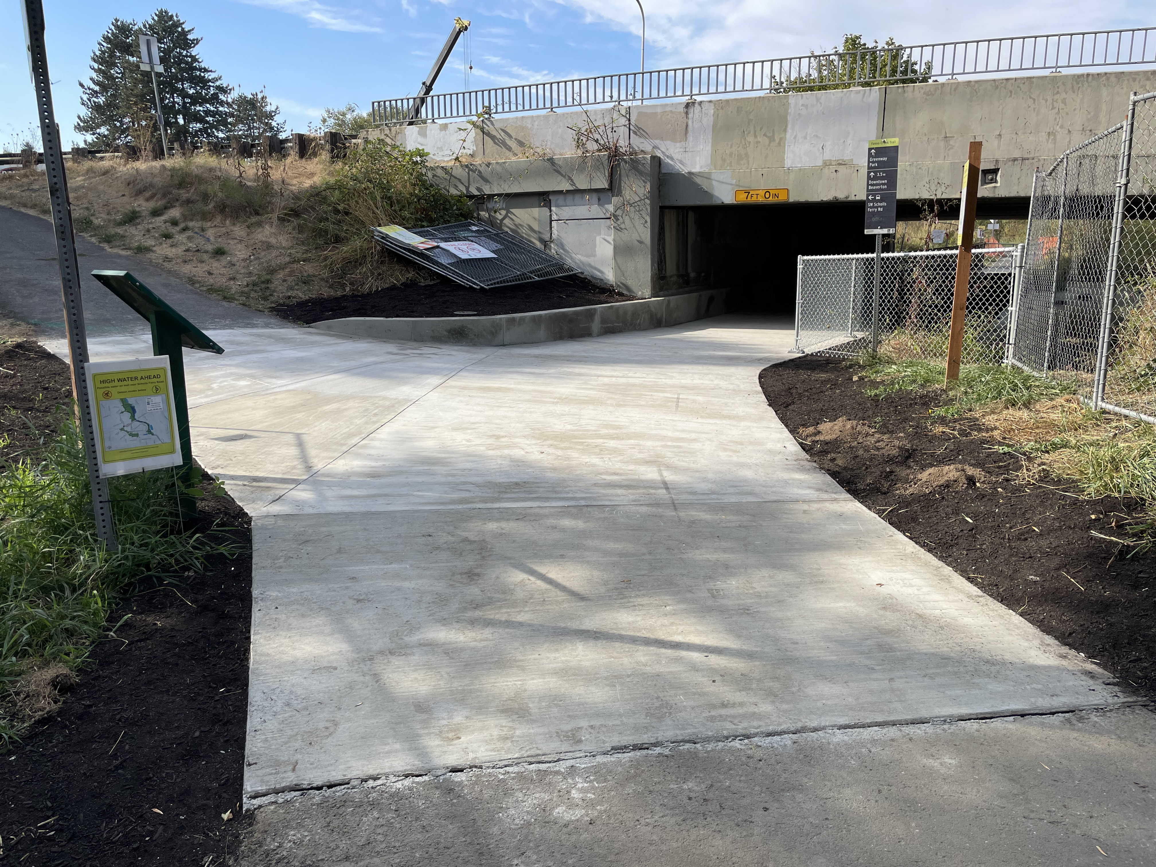 A concrete trail runs under an overpass. Some signs of construction, such as fencing, are visible.