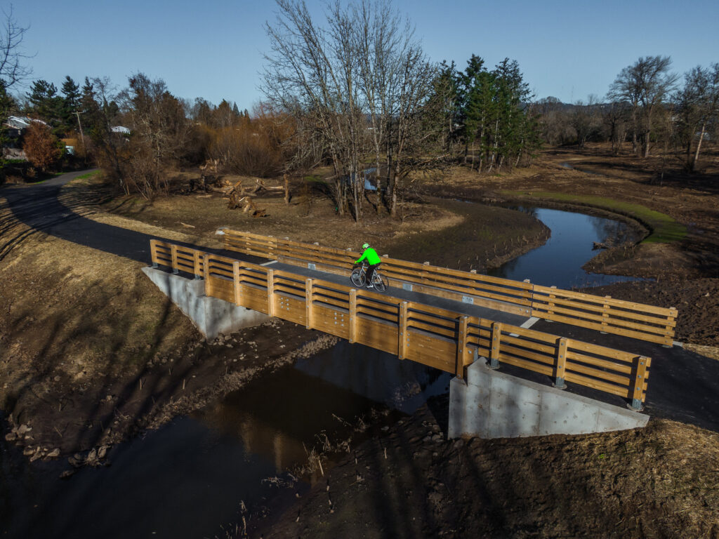 A community member wearing a bright yellow shirt and helmet rides a bike across a bridge at Fanno Creek