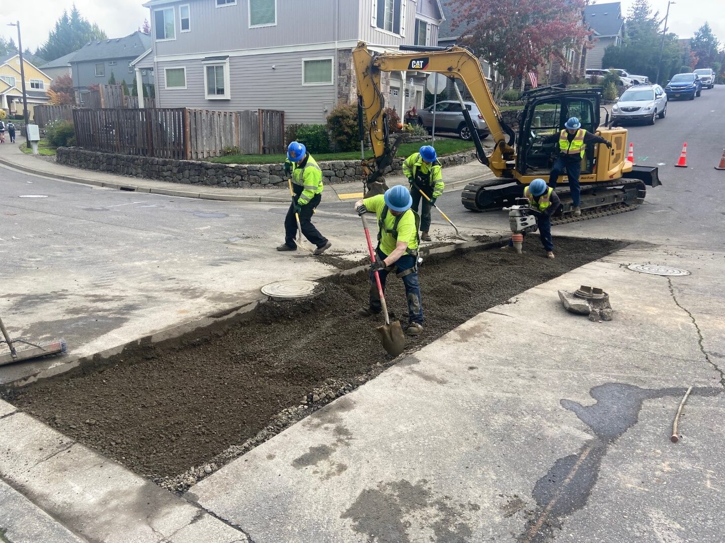 Workers fill a trench in the street where pipe work was completed. A piece of heavy machinery, houses, and cars are in the background.