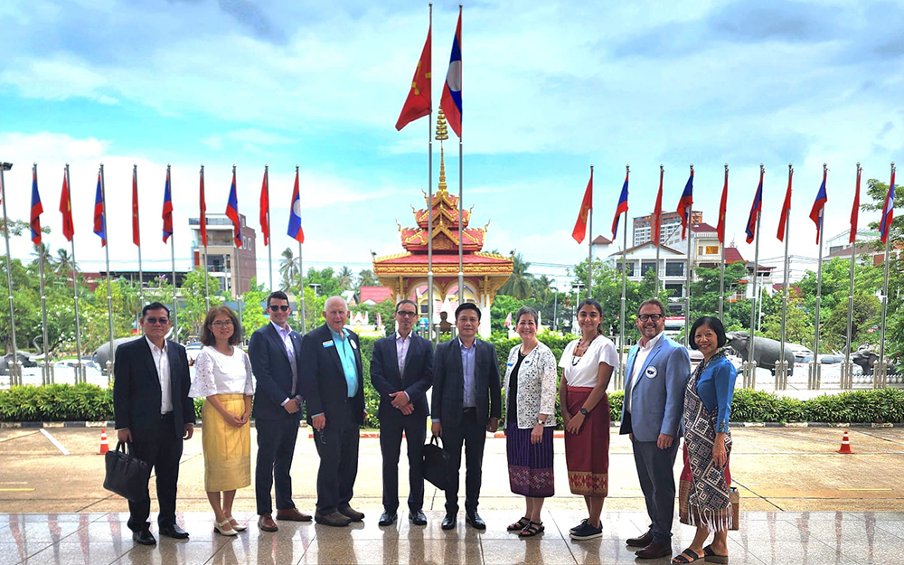 Ten people stand in a line posing for a photo. In the background is a row of flags and a city scape with buildings and trees.