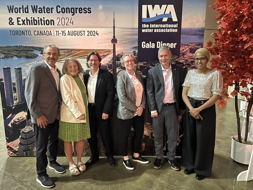 Five people stand in a line posing in from of a sign for the International Water Association World Water Congress & Exhibition 2024 in Toronto, Canada