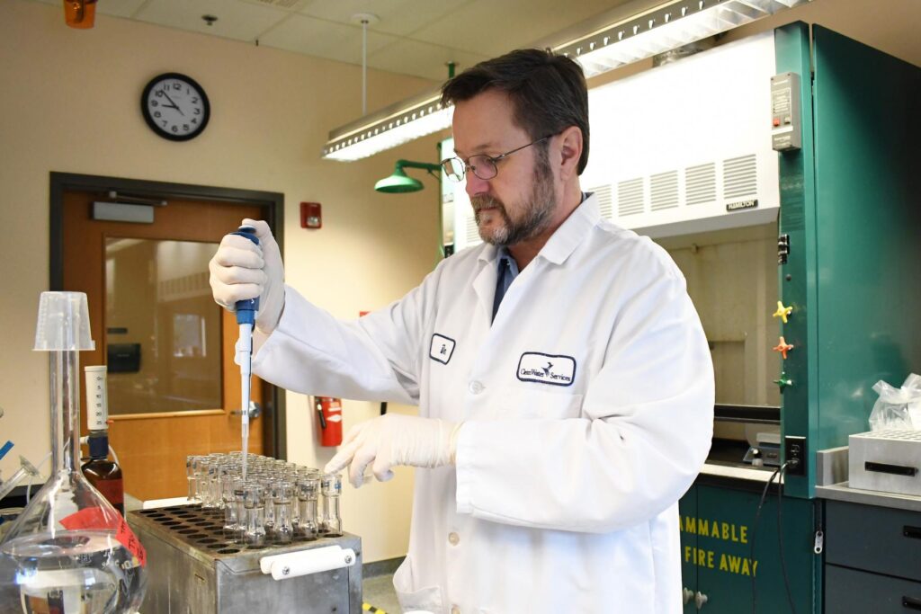 In a laboratory, a chemist in a white lab coat and latex gloves pushes the button at the top of a device that delivers a water sample into one of several glass vials.