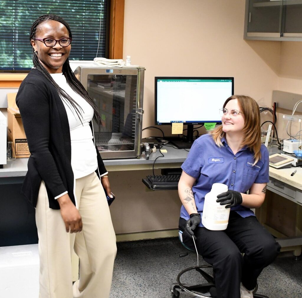 A woman smiles as she looks at the camera, as another woman sitting in a chair looks at her, also smiling, while holding a water sample jug. Laboratory equipment and a computer are on the counter behind them.
